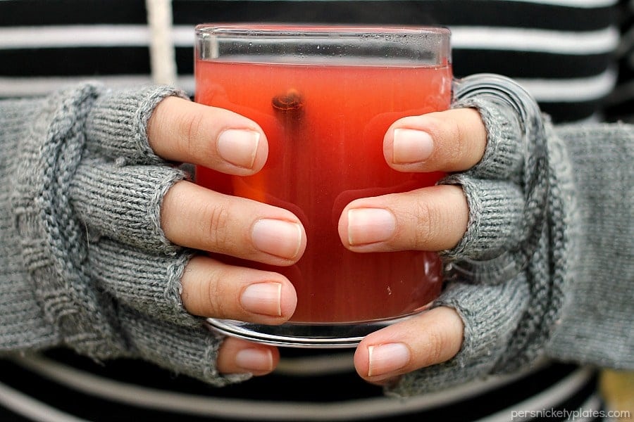 hands wearing fingerless gloves holding a glass mug of cider. 
