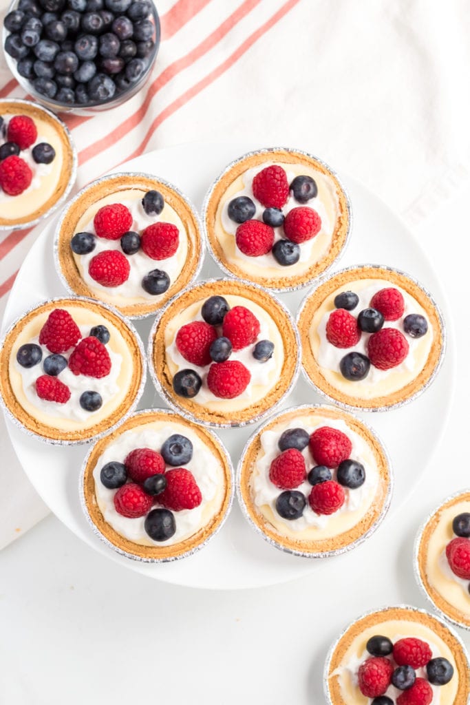 7 mini cheesecakes topped with raspberries & blueberries on a plate.