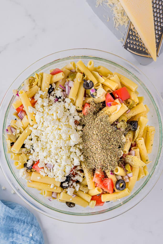 overhead shot of seasonings sprinkled on pasta salad in a mixing bowl.