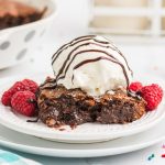 chocolate fudge pie on a white plate topped with vanilla ice cream, drizzled with fudge, and raspberries.