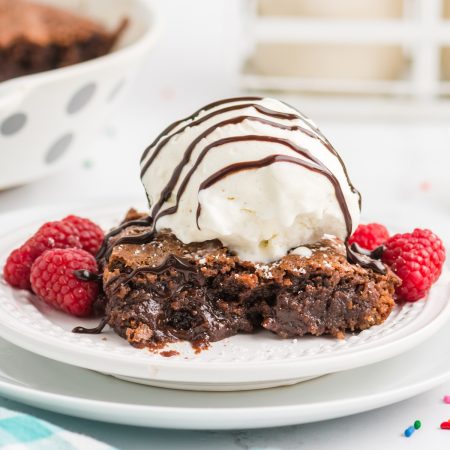 chocolate fudge pie on a white plate topped with vanilla ice cream, drizzled with fudge, and raspberries.