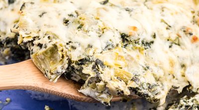 wooden spoon scooping spinach artichoke chicken casserole from a blue glass dish.