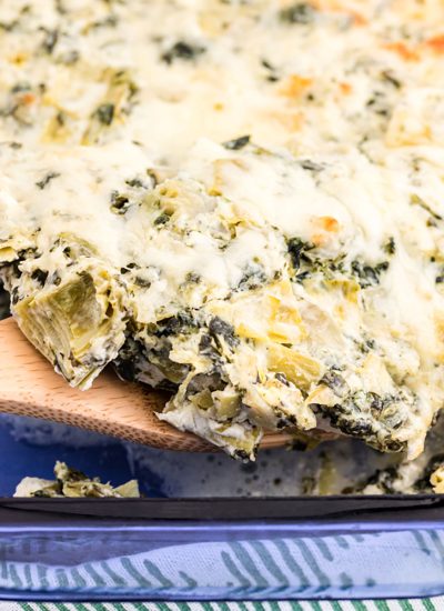 wooden spoon scooping spinach artichoke chicken casserole from a blue glass dish.