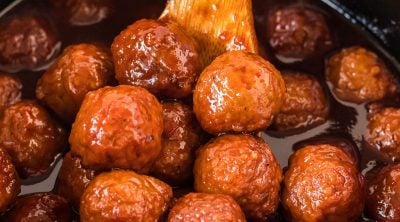 wooden spoon scooping grape jelly meatballs from a crockpot.