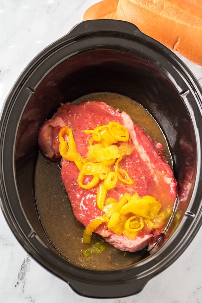 overhead shot of a roast beef in a slow cooker topped with peppers.