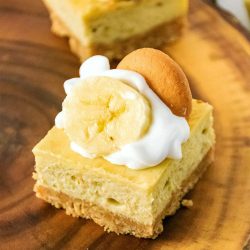 a banana pudding cheesecake bar on a wood plate topped with whipped cream, banana slice, and a vanilla wafer.