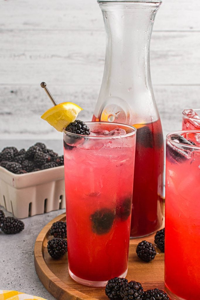 glass of blackberry lemonade with lemon garnish in front of carafe.