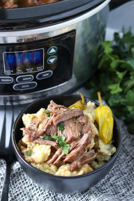 pot roast in a bowl over potatoes in front of a slow cooker.