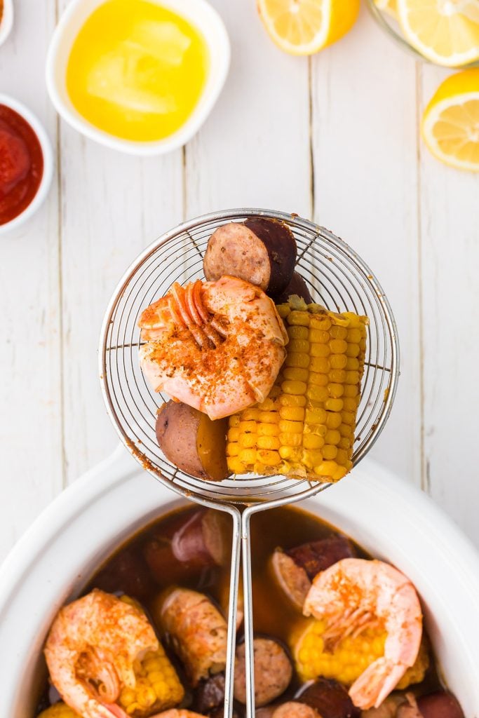 slotted spoon lifting shrimp and corn from a slow cooker.
