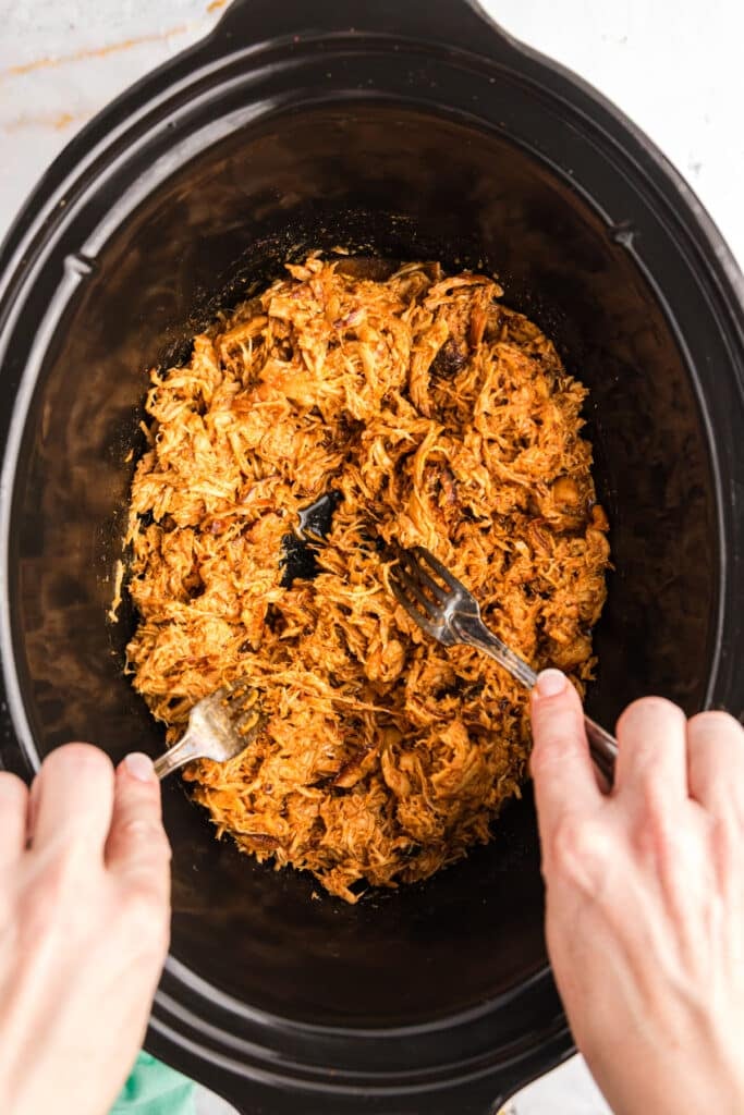 two forks shredding chicken in a slow cooker.