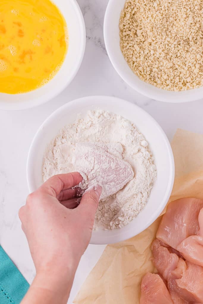hand dipping chicken into flour.