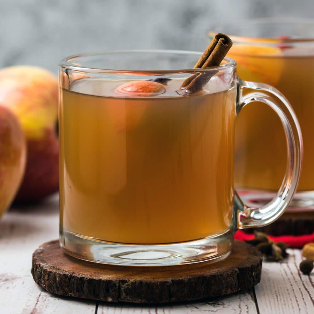 closeup of a clear glass mug of apple cider with a cinnamon stick.