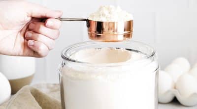 full measuring cup of flour being held over a glass jar of flour.