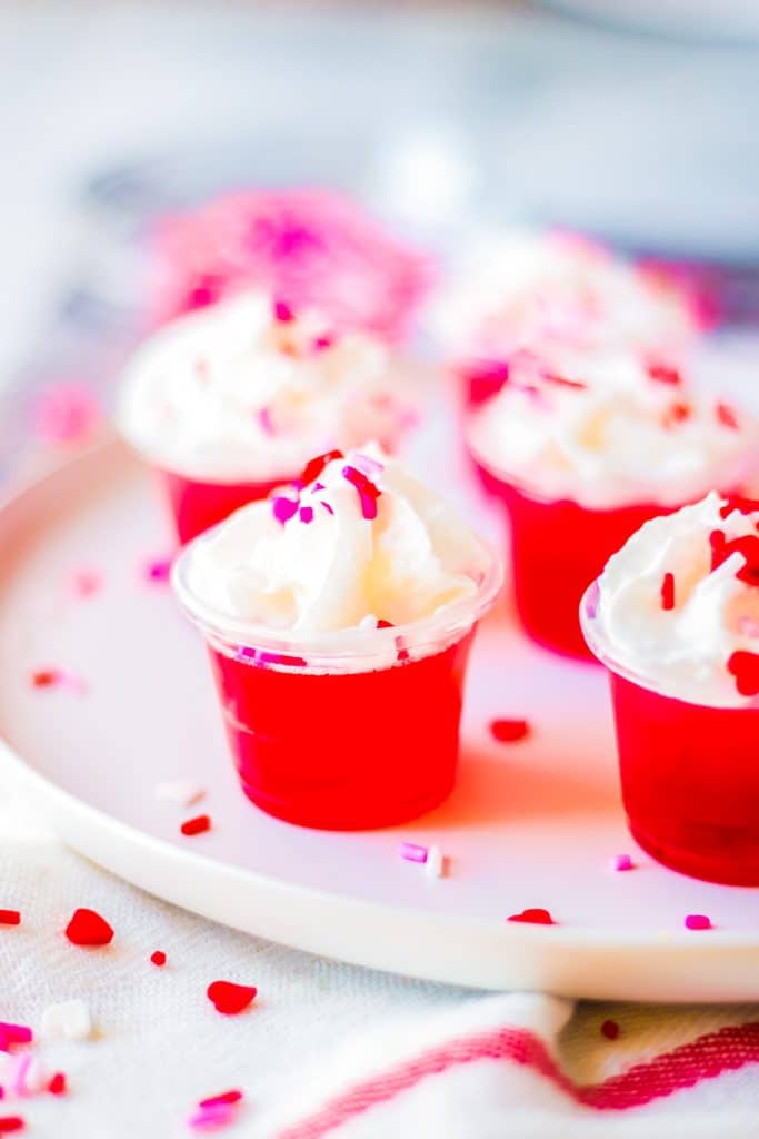valentine's day jello shots on a white plate with whipped cream & sprinkles.