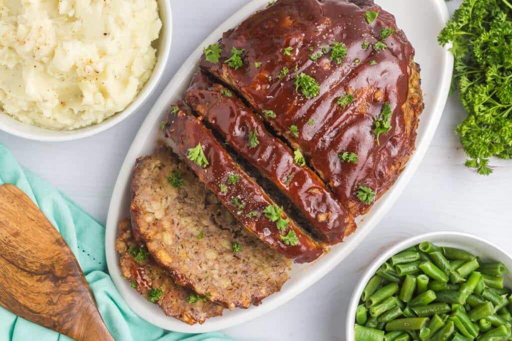 overhead shot of sliced meatloaf on a platter with mashed potatoes & green beans.