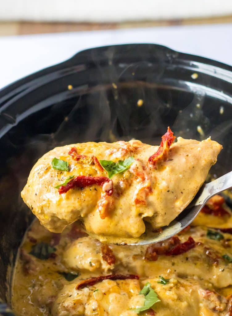 spoon lifting a chicken breast with sun dried tomatoes & basil from a crockpot.