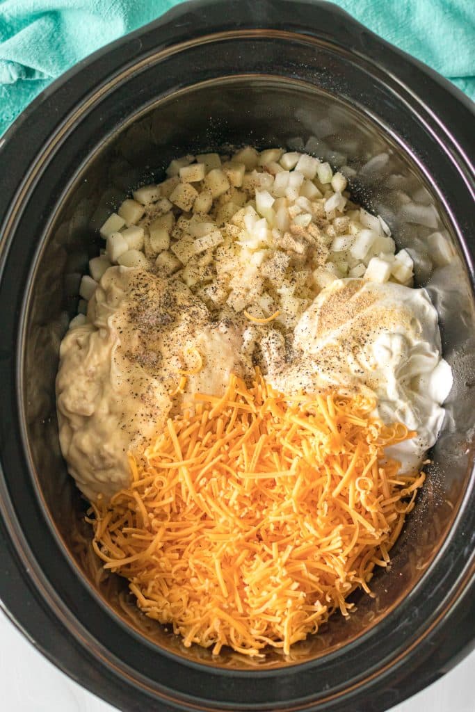 overhead shot of diced potatoes, shredded cheese, sour cream, and seasonings in a black slow cooker.