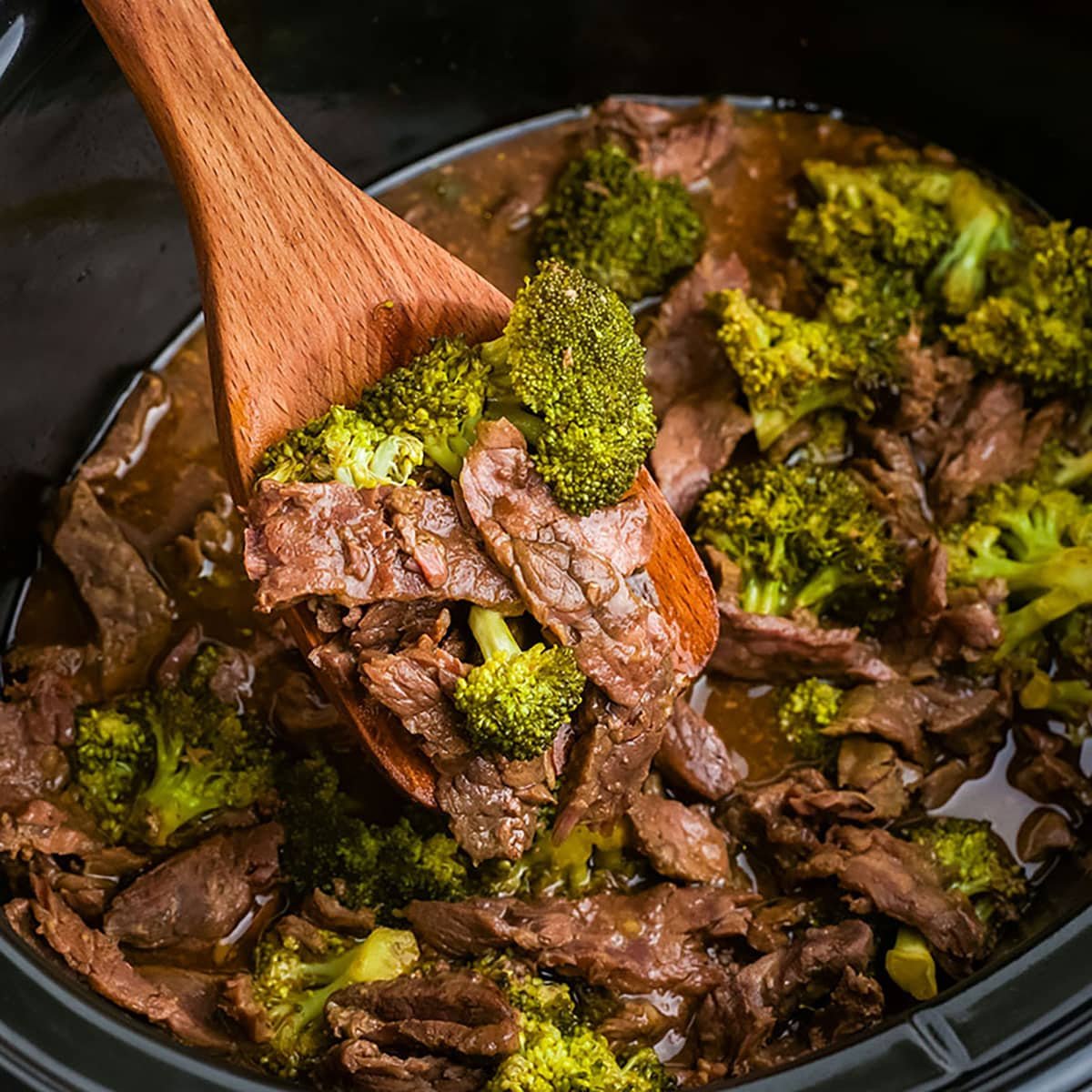 wood spoon lifting a scoop of beef & broccoli from a slow cooker.