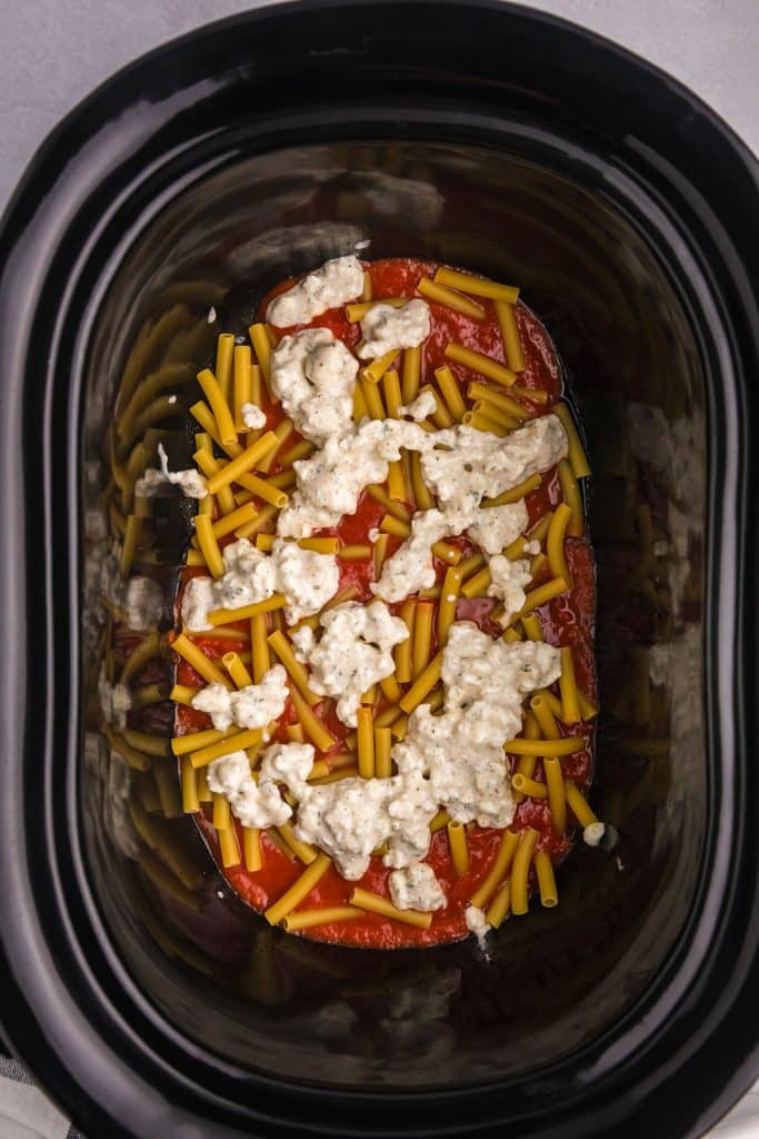 cheese mixture spread on uncooked noodles in a black crockpot.
