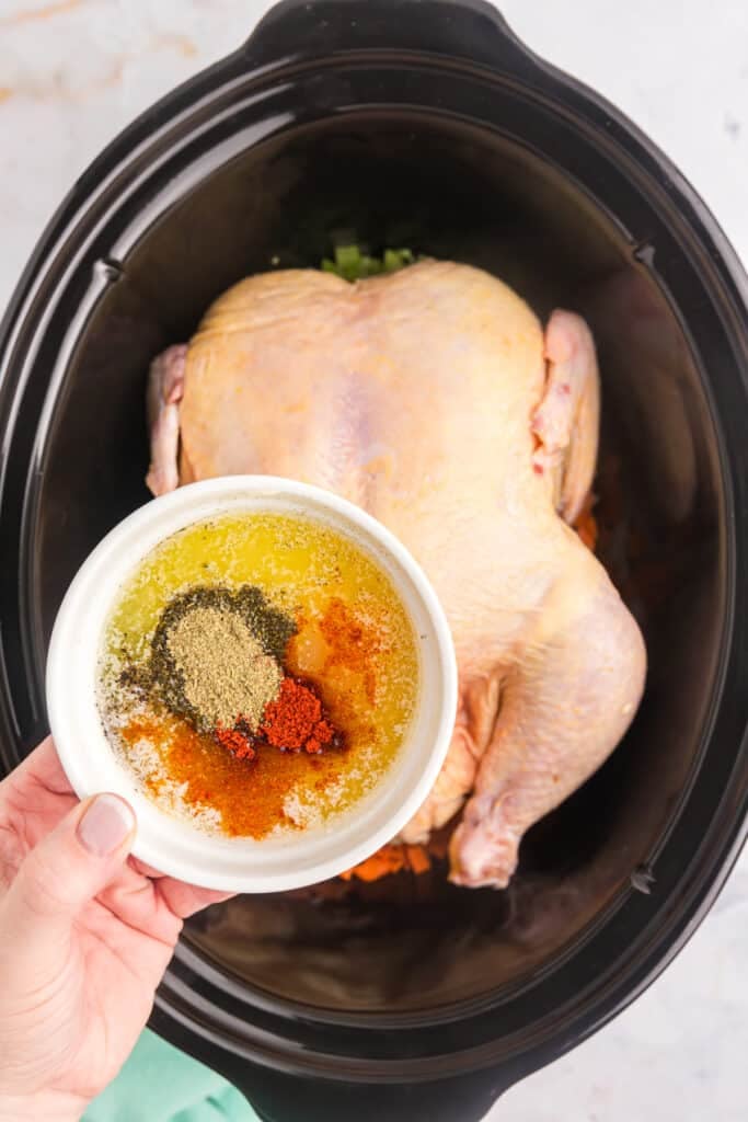 hand holding a bowl of butter and seasonings over a whole chicken.