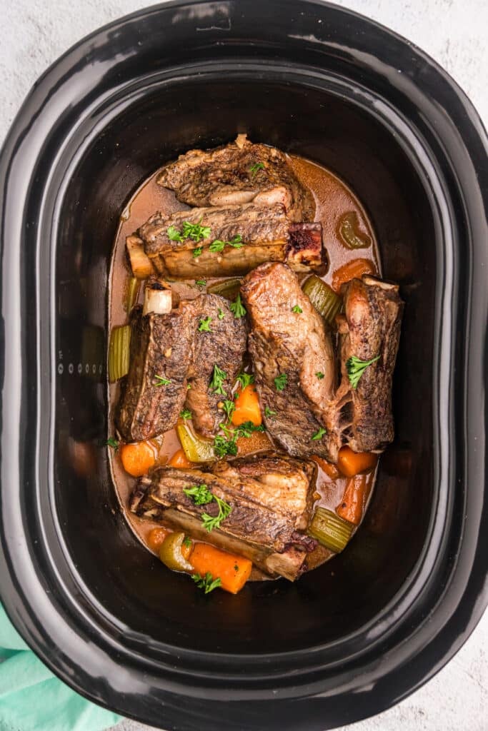 overhead shot of beef short ribs, carrots, and celery in a black slow cooker.
