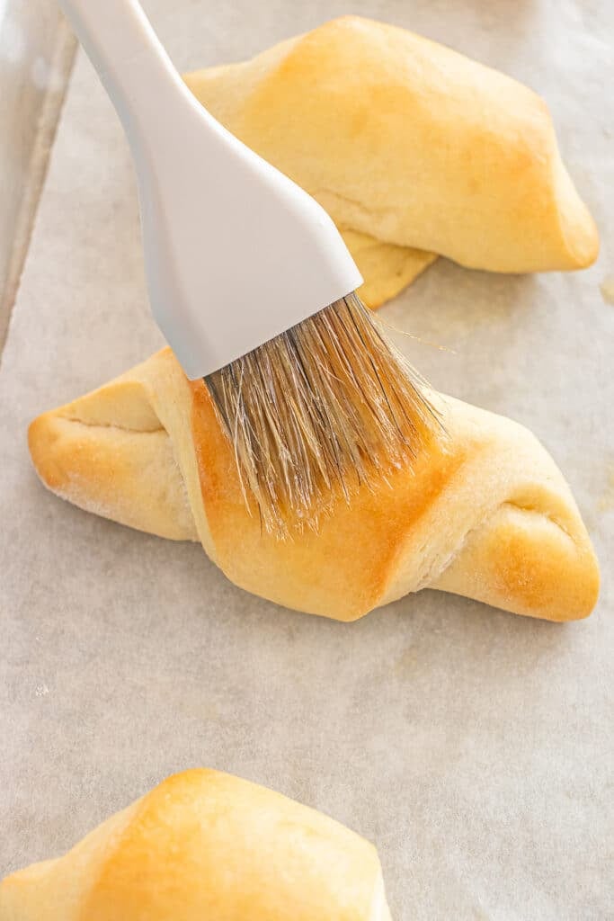 A close-up of a pastry brush spreading butter on a baked crescent roll.