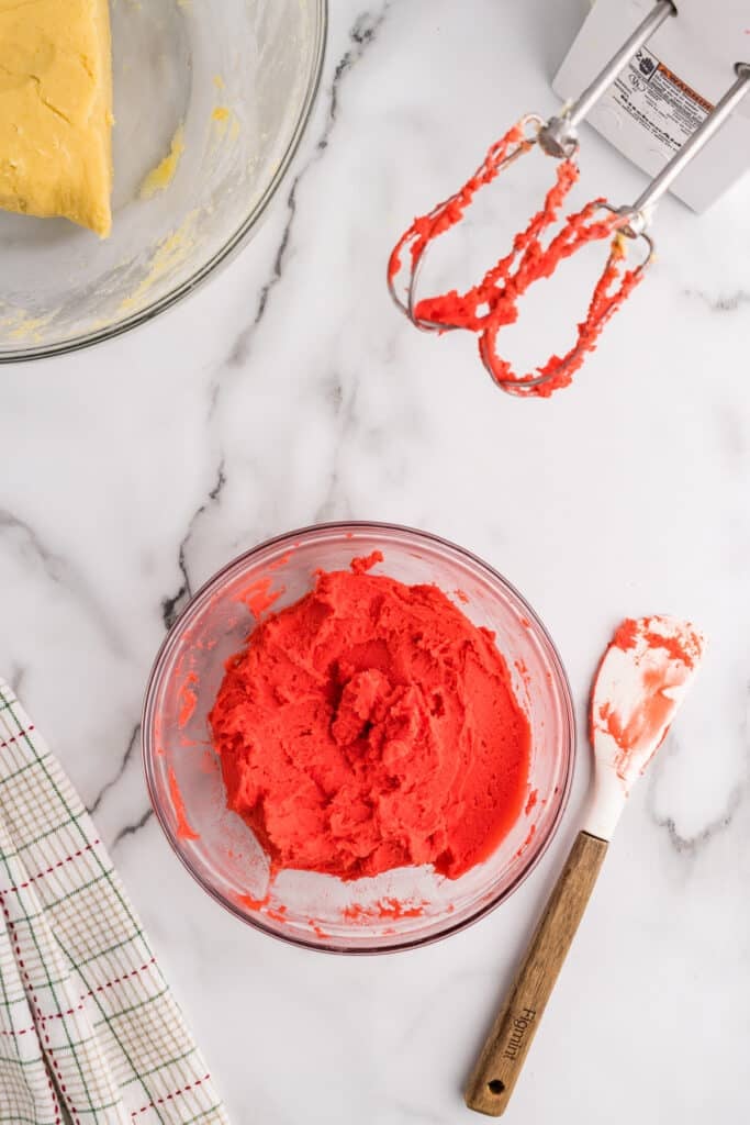 Red cookie dough in a glass bowl with mixer beaters nearby.
