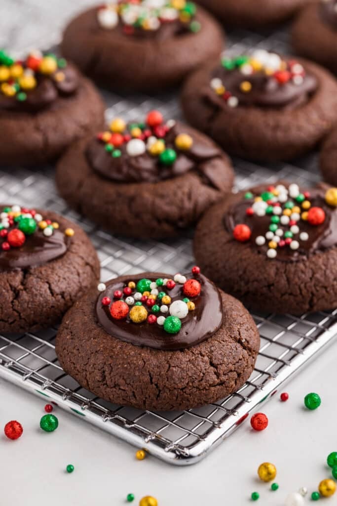 A cooling rack lined with chocolate thumbprint cookies, each with a chocolate ganache center and colorful round sprinkles.