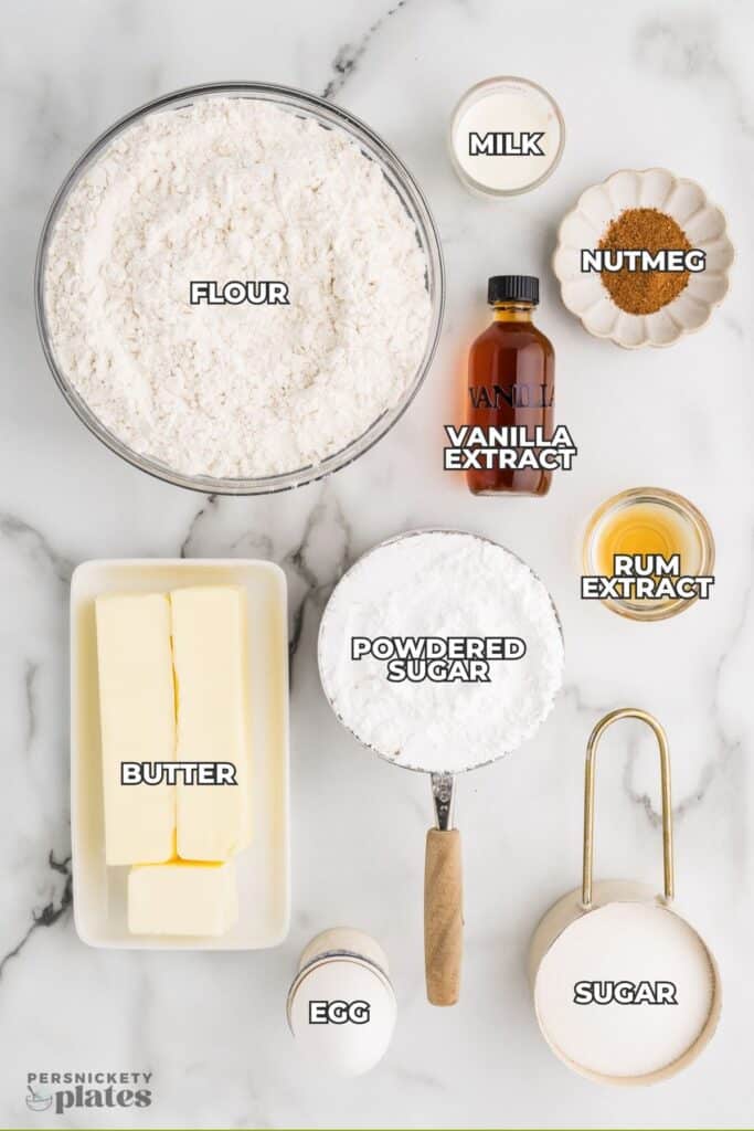 A flatlay of baking ingredients labeled with text, including flour, butter, sugar, egg, spices, and extracts.