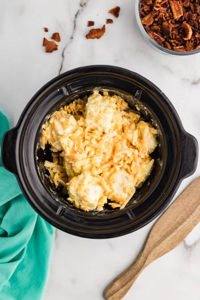 A slow cooker containing large chunks of cream cheese coated with shredded cheese before melting, shown before the ingredients are mixed together.