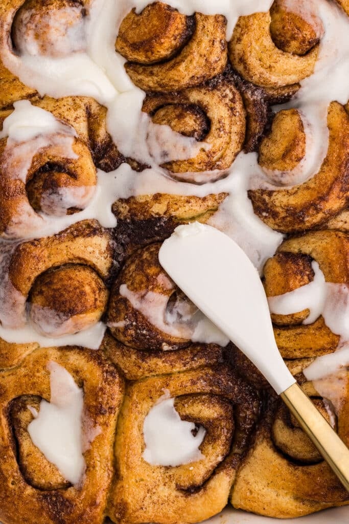 Cinnamon rolls with icing being spread using a spatula.