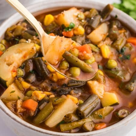 Spoon lifting chunky vegetable soup with potatoes, green beans, peas, corn, and carrots in a broth.