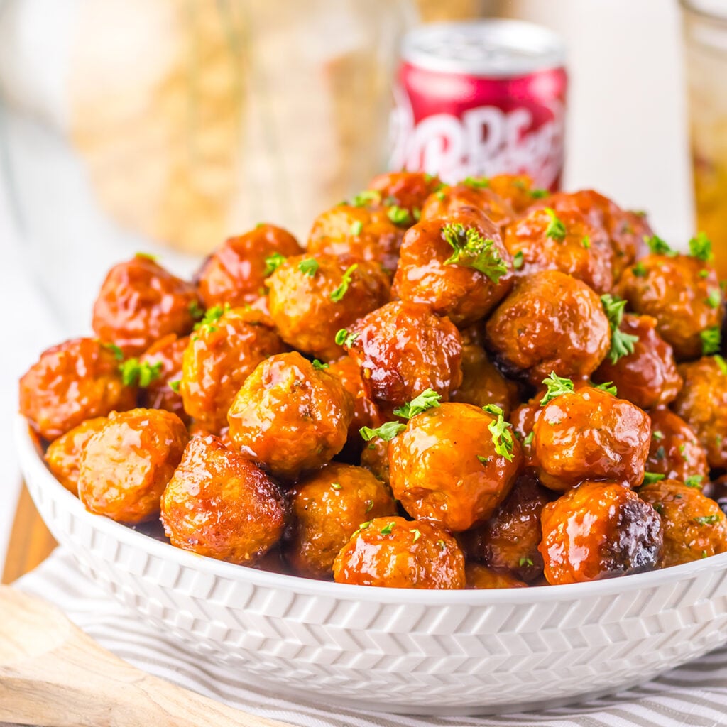 A white serving bowl filled with fully glazed meatballs and garnished with parsley.