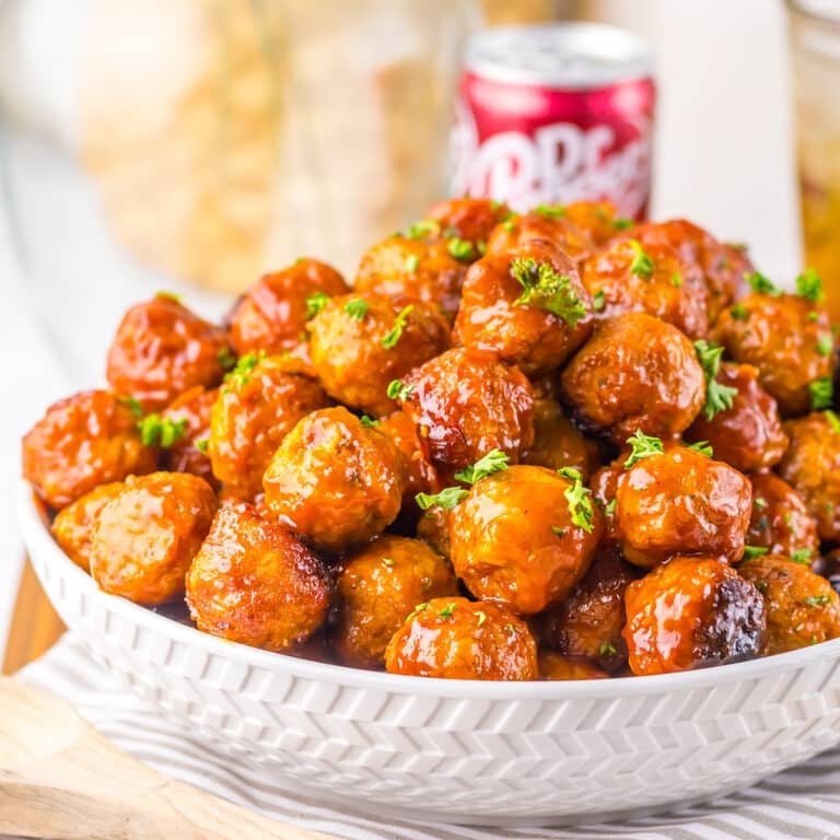 A white serving bowl filled with fully glazed meatballs and garnished with parsley.