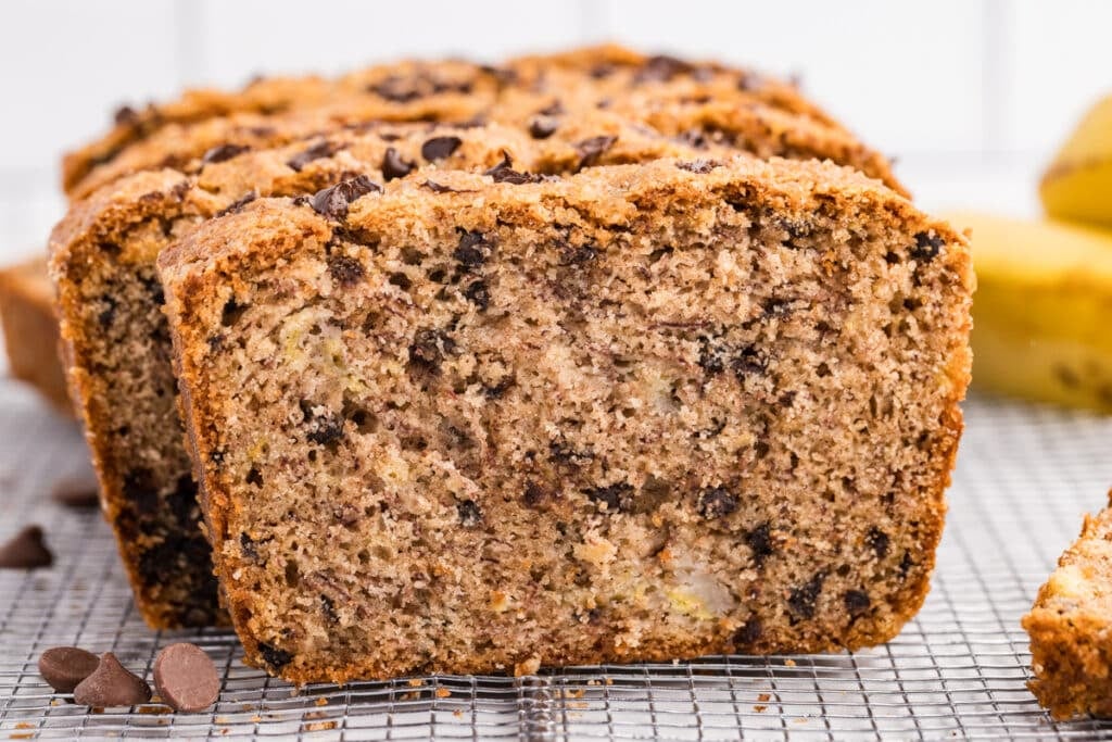 Close-up of a sliced loaf of chocolate chip banana bread on a wire cooling rack with bananas in the background.