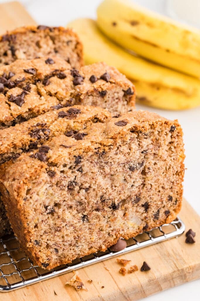Sliced chocolate chip banana bread loaf on a cooling rack with bananas in the background.