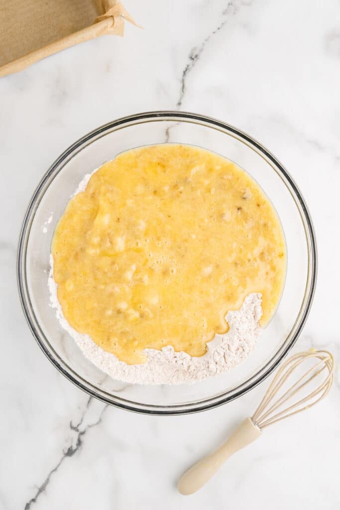Wet banana mixture poured over flour in a glass mixing bowl with a whisk beside it.