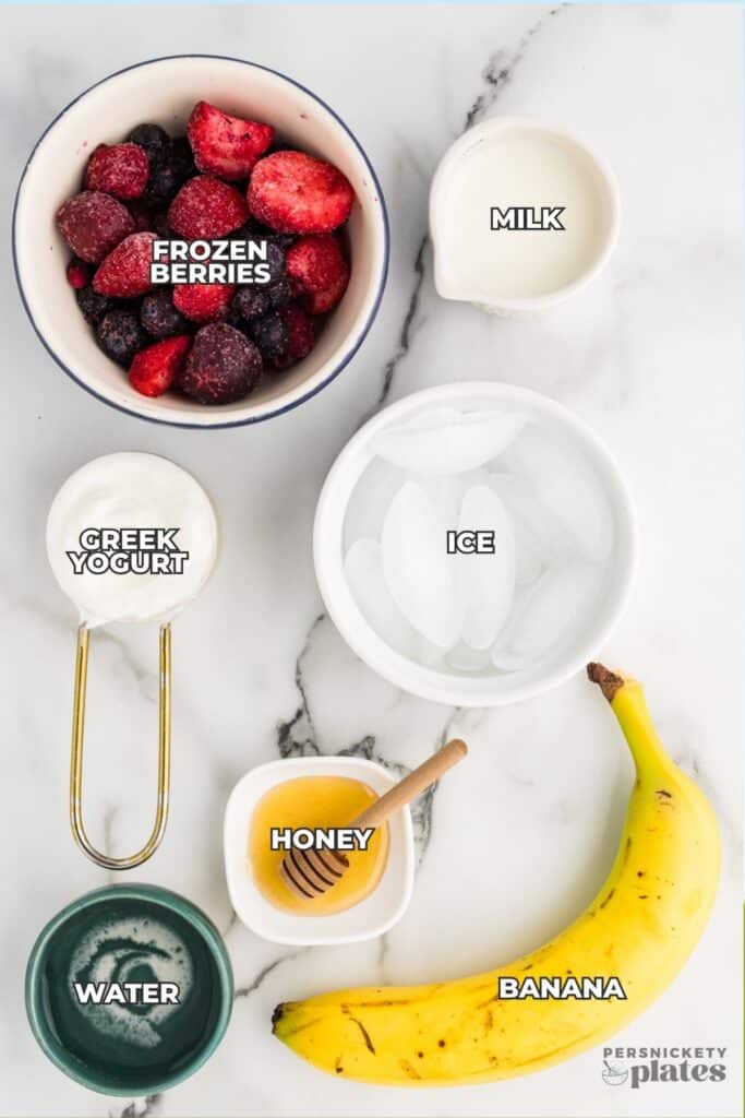 Overhead view of smoothie ingredients including frozen berries, banana, milk, Greek yogurt, ice, honey, and water labeled on a marble surface.