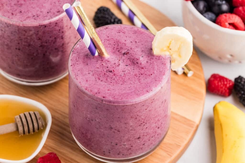 Glass of mixed berry smoothie with a banana slice garnish and paper straws on a wooden board with berries and honey nearby.