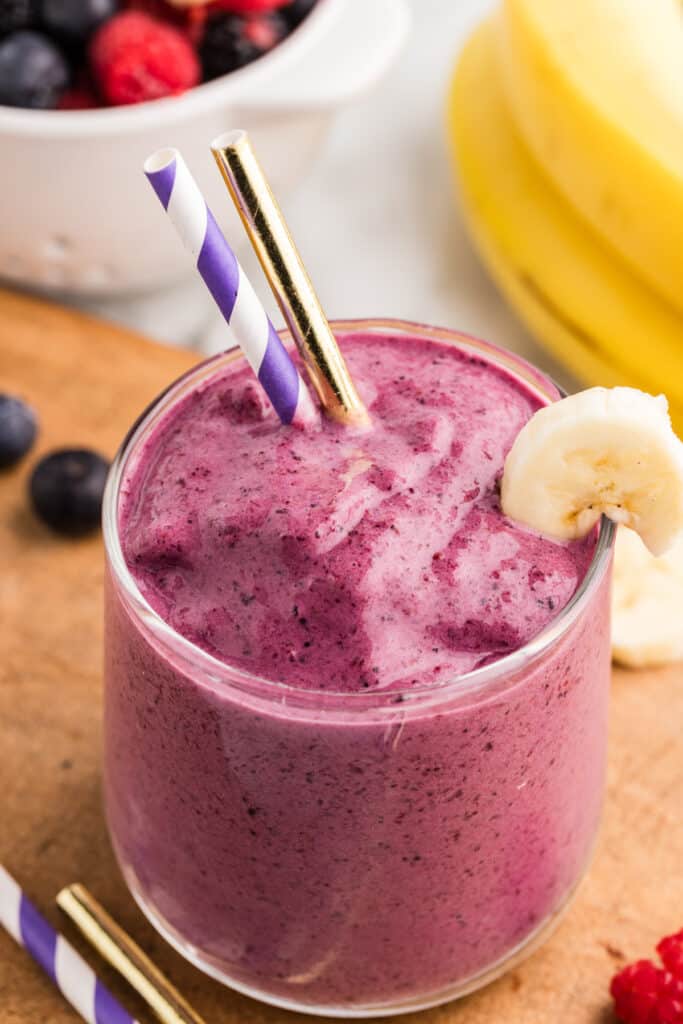 Glass of thick mixed berry smoothie with a banana slice garnish and two straws on a wooden board.