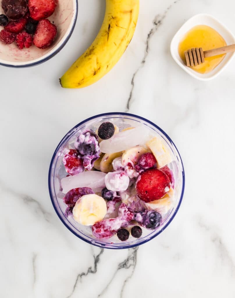 Frozen mixed berries, banana slices, yogurt, and ice in a blender cup on a marble countertop.