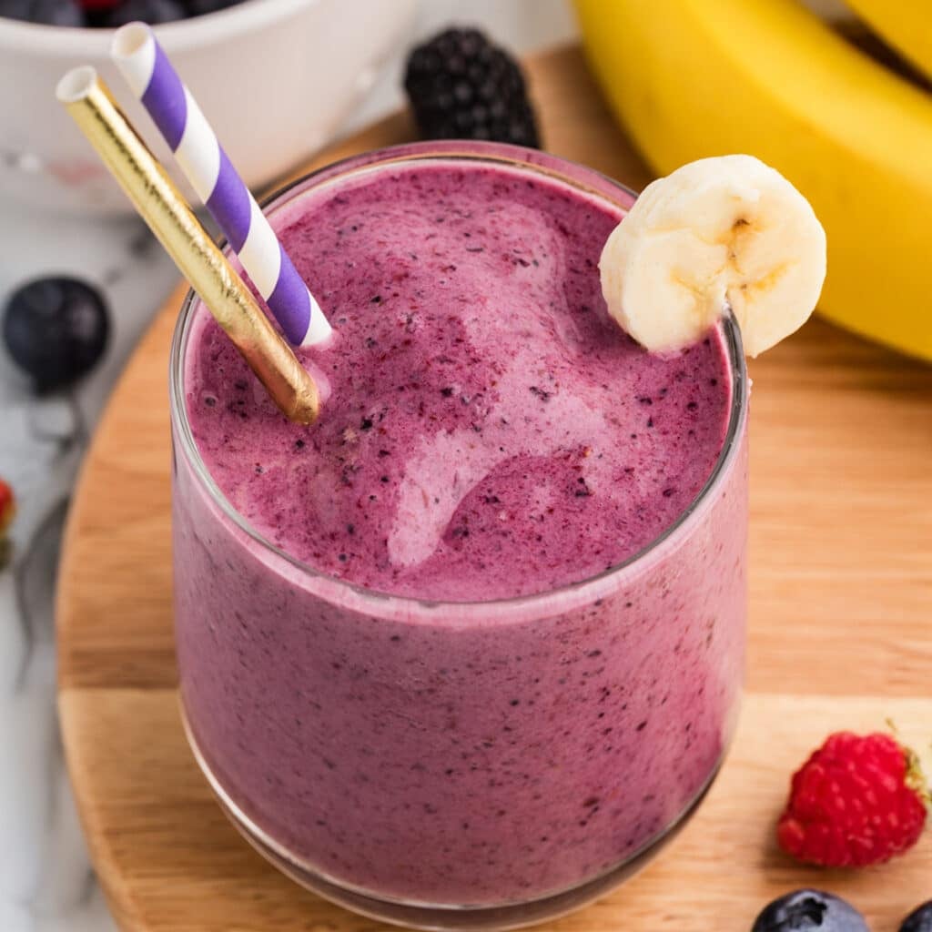 Glass of thick mixed berry smoothie topped with foam, garnished with a banana slice and paper straws.