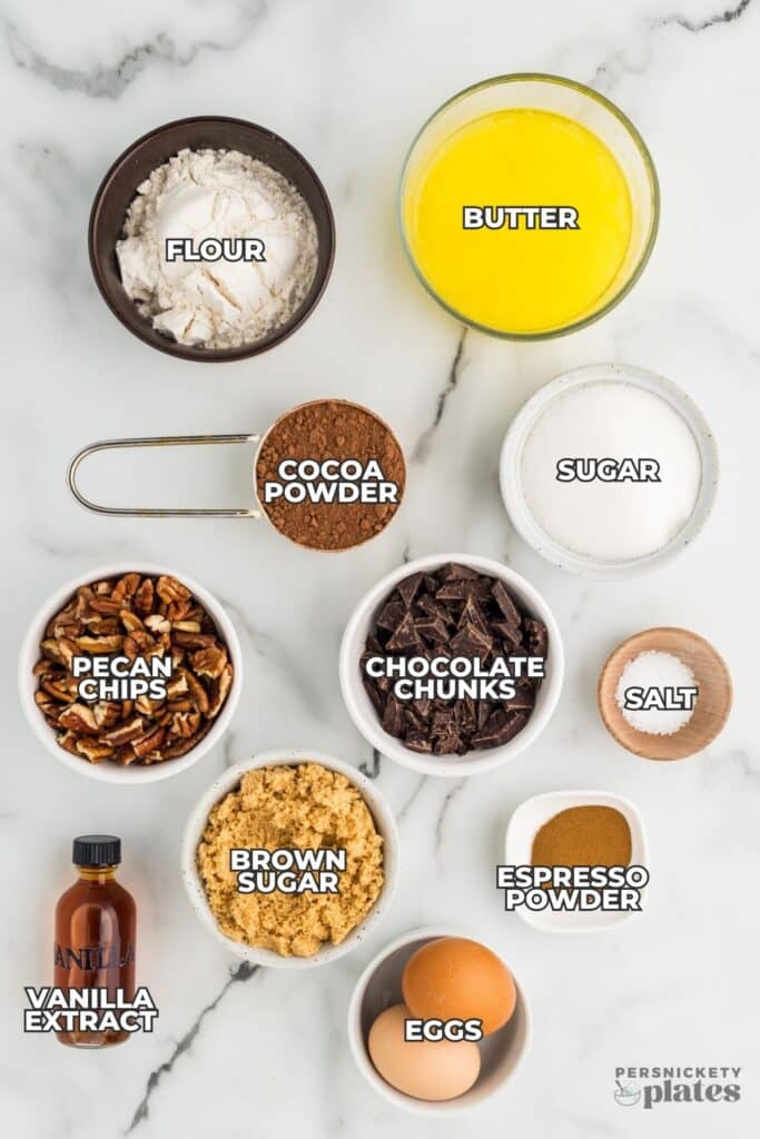 Overhead layout of brownie ingredients labeled flour, butter, cocoa powder, sugar, pecan chips, chocolate chunks, salt, brown sugar, espresso powder, vanilla extract, and eggs.