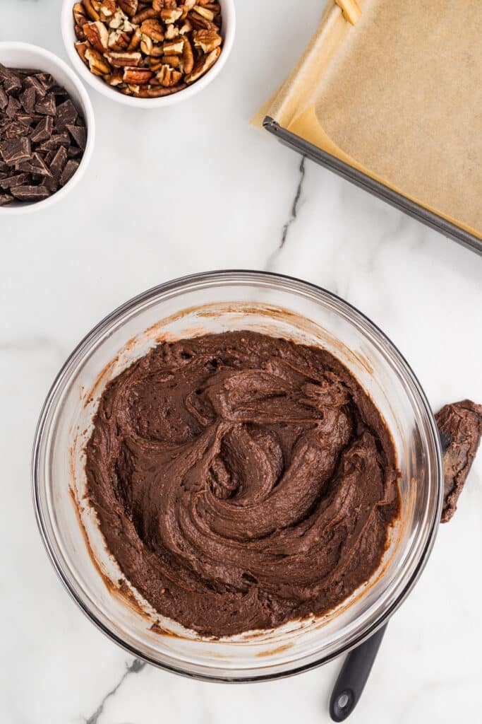 Glass bowl filled with thick chocolate brownie batter on a marble countertop with bowls of pecans and chocolate nearby.