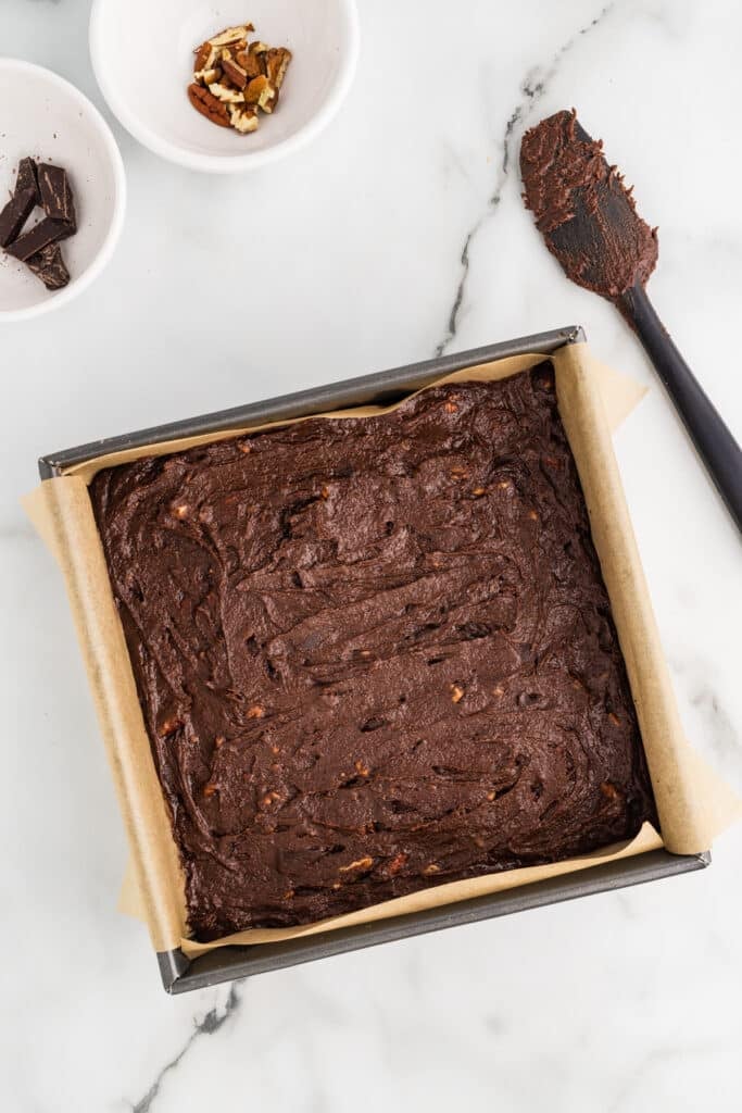 Square pan of chocolate brownie batter lined with parchment paper on a marble countertop.