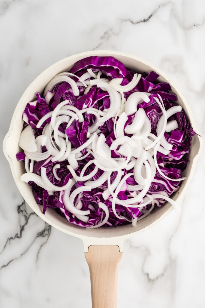 Sliced red cabbage and white onions in a skillet on a marble surface.