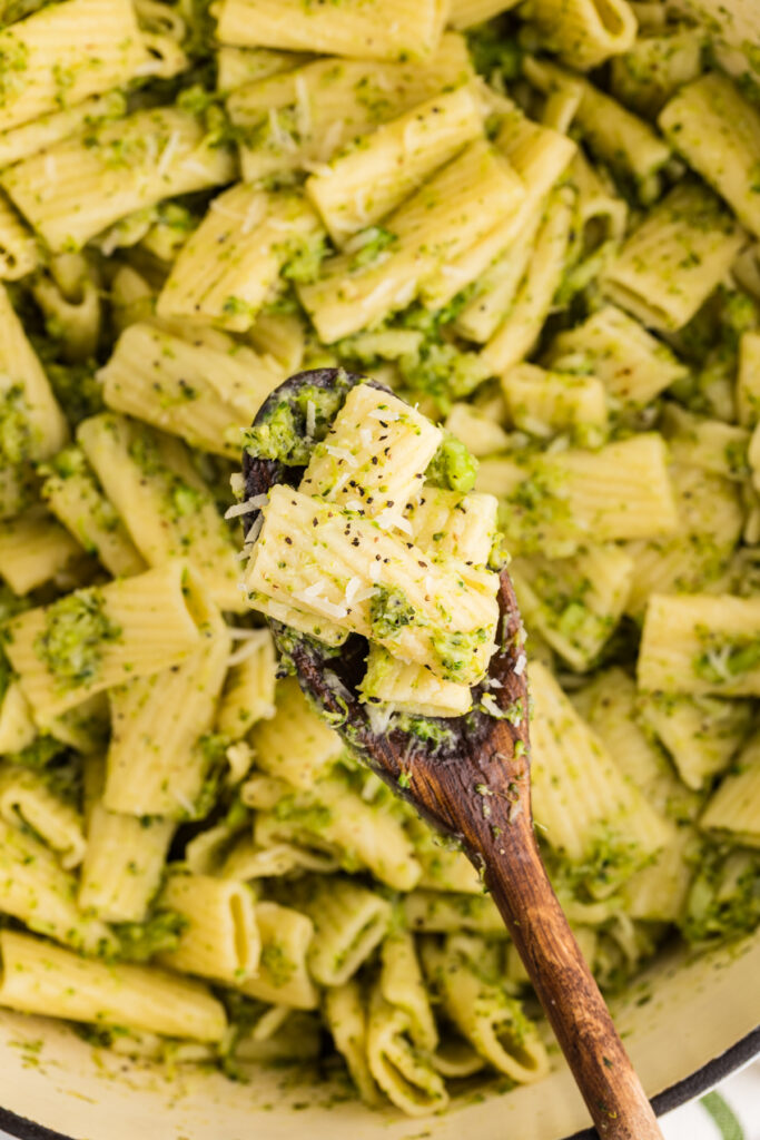 a wooden spoon lifting rigatoni pasta coated in a green broccoli sauce with grated cheese and black pepper, held above a pot filled with more pasta.