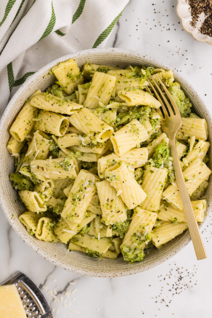 a bowl filled with rigatoni pasta and chopped broccoli in a light green sauce, topped with grated cheese, with a gold fork resting inside the bowl.