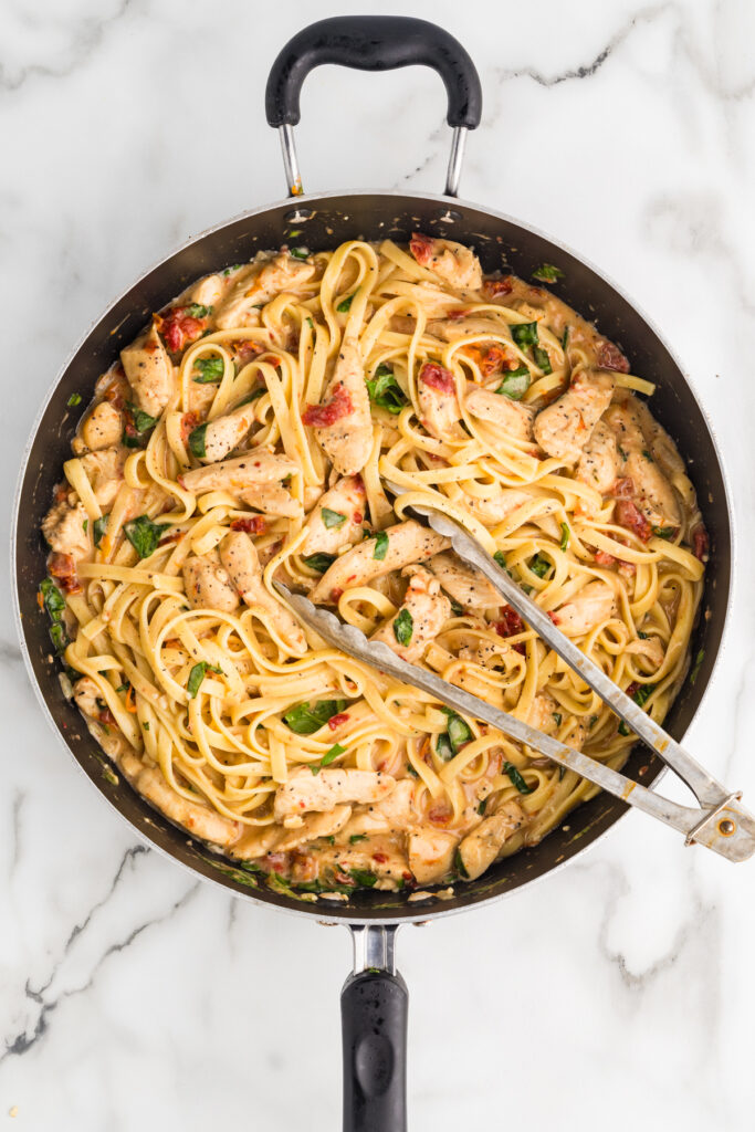 A black skillet filled with cooked pasta, sliced chicken, sun-dried tomatoes, and green herbs tossed together.