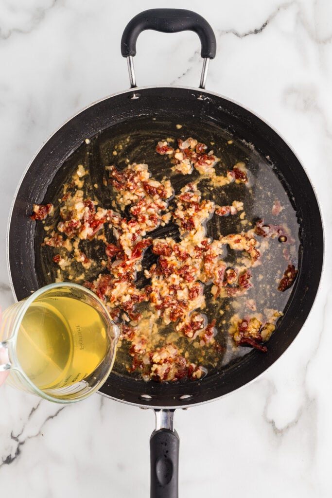 Broth being poured into a skillet with sun-dried tomatoes and garlic